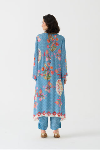 A woman viewed from behind wearing a light blue kurta with colorful floral embroidery and matching blue pants, standing against a plain white studio background.